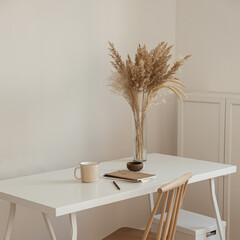 Women's home office workspace. Freelance work, business workplace. Light aesthetic hygge space with wooden chair, table, reed pampas grass bouquet, mug, notebook. Modern interior design.