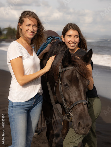 Two women hugging brown horse. Horse riding on the beach. Human and animals relationship. Nature concept. Copy space. Bali, Indonesia © Olga