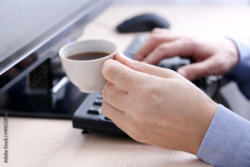 Man drinks coffee sitting at PC keyboard. Cup of hot drink in male hand  break during work in office