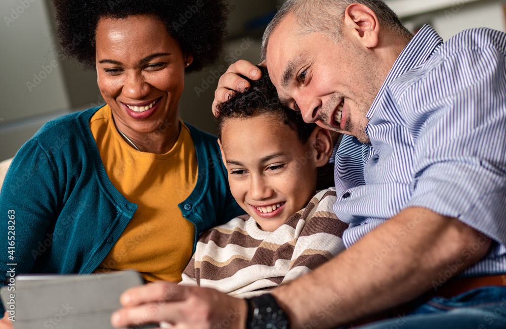 Smiling mixed race family enjoying time at home sitting on sofa in living room looking at tablet.