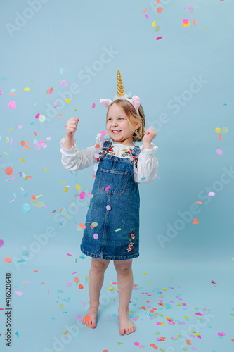Happy little blonde girl in unicorn head band and jeans overalls stretching over blue background.