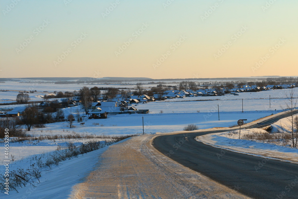 An asphalt road in the middle of blue snow fields in golden from sunset superhorns. In the distance there is a winter village with houses and trees along the street. Winter rural bluish-gold landscape