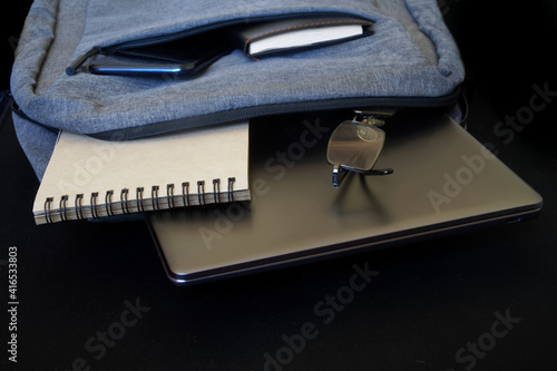 A small gray city backpack with a laptop or ultrabook, glasses, a smartphone and a notepad lies on a black background. Attributes of an urban traveler, programmer or student. Free space