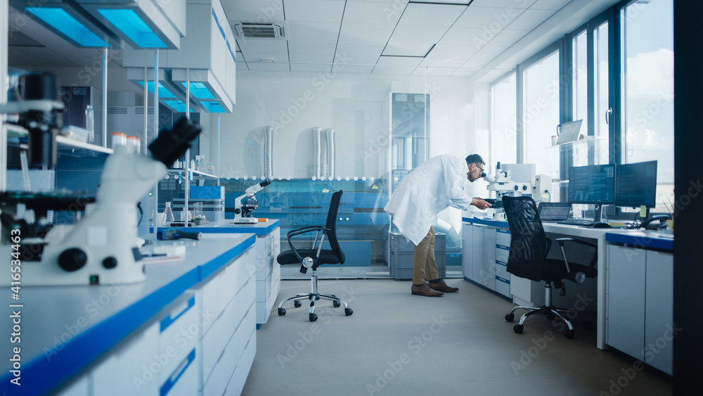 Modern Medical Research Laboratory with Male Scientist Looking Under Microscope, Analysing Microbiological Sample. Advanced Scientific Lab for Drugs, Vaccine Development, Full of High-Tech Equipment Modern Medical Research Laboratory with Male Scientist Looking Under Microscope, Analysing Microbiological Sample. Advanced Scientific Lab for Drugs, Vaccine Development, Full of High-Tech Equipment