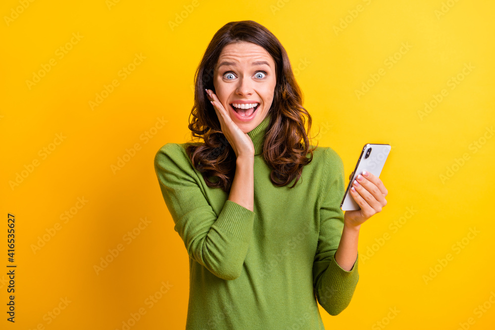 Portrait of young shocked amazed happy surprised woman use smartphone hold hand cheek isolated on yellow color background