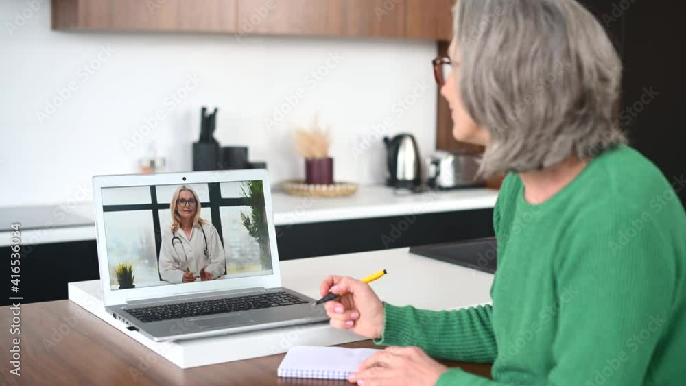 Mature senior lady talking to a doctor online from home, having a virtual medical check-up with the medical specialist on the laptop, taking a notes. Telemedicine concepts Mature senior lady talking to a doctor online from home, having a virtual medical check-up with the medical specialist on the laptop, taking a notes. Telemedicine concepts