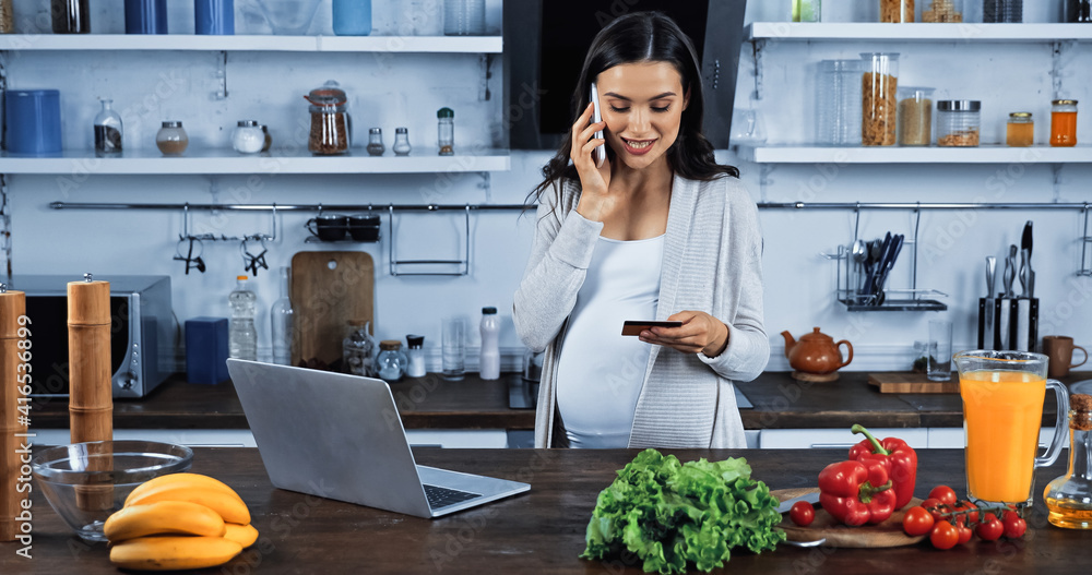 Smiling pregnant woman talking on smartphone while holding credit card near food in kitchen Smiling pregnant woman talking on smartphone while holding credit card near food in kitchen