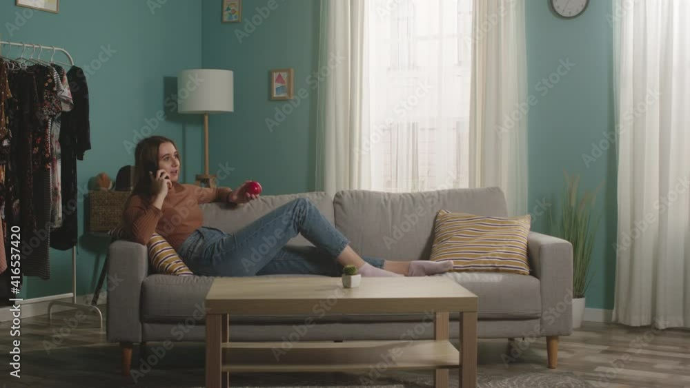 Young lady in blue jeans and a brown sweater lies on a beige sofa on the background of the window, holding a red apple and talking on the phone. General plan. Young lady in blue jeans and a brown sweater lies on a beige sofa on the background of the window, holding a red apple and talking on the phone. General plan.