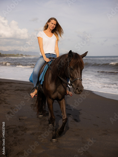 Beautiful woman riding horse on the beach. Outdoor activities. Caucasian woman wearing jeans and white T-shirt. Traveling concept. Cloudy sky. Bali, Indonesia