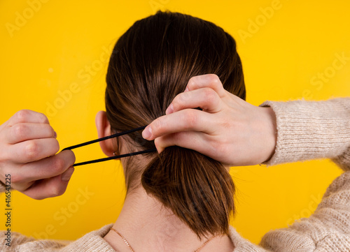 Back view shot of a fair-haired lady. There's a black rubber band on her hair with a black fake hair braid. The photo was taken against a yellow background.