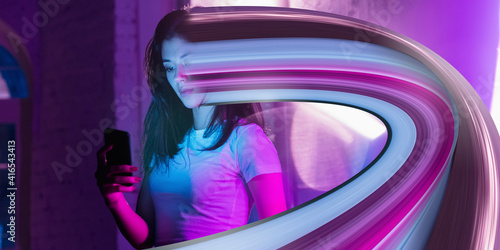 Cinematic portrait of stylish woman in neon lighted interior using a smartphone. The face is smeared, sucked, absorbed into the phone. Concept of social network dependency, phone addiction.