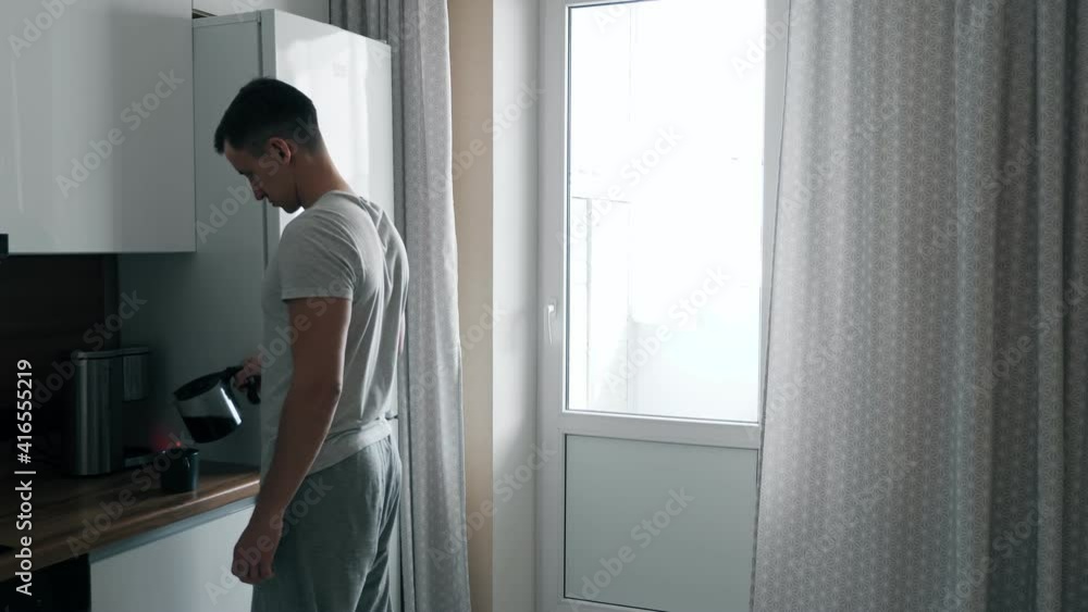Handsome young man pours coffee from coffee maker and walks up to window in kitchen. Handsome young man pours coffee from coffee maker and walks up to window in kitchen.