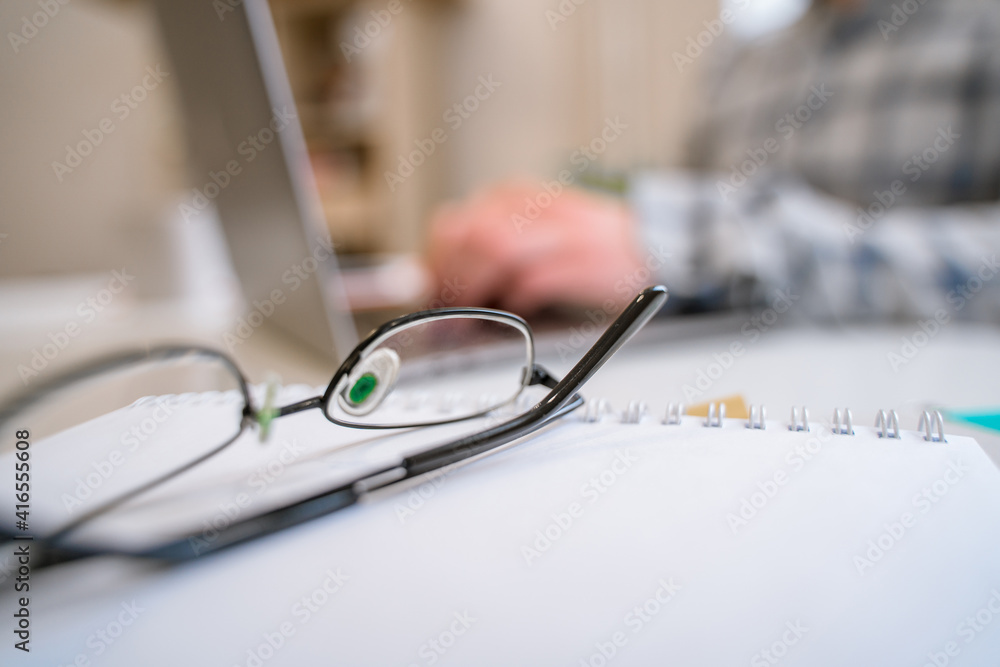 A white desk with a focus on glasses, a man working on a laptop in the background A white desk with a focus on glasses, a man working on a laptop in the background