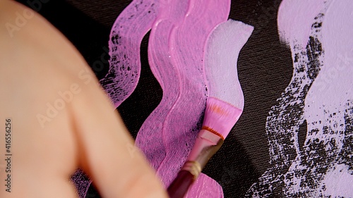 Artist's hand holds a paintbrush and draws a wavy line of pink or lilac on the black canvas. Close view. Pink color of oil paint or watercolor. Acrylic drawing concept, artist work