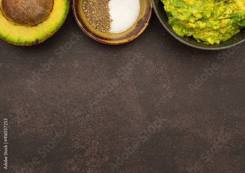 Top view of fresh guacamole on a dish placed on a dark gray stone background with ingredients for homemade guacamole: avocado, salt, and pepper. Concept of traditional Mexican preparation