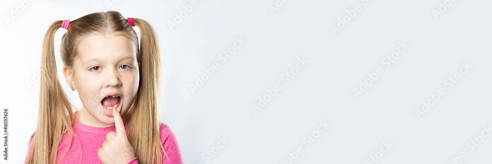 Little caucasian girl with long blond hair in pink clothes shows a milk tooth in her mouth to be pulled out on a light background Little caucasian girl with long blond hair in pink clothes shows a milk tooth in her mouth to be pulled out on a light background