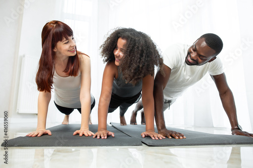 Happy multiracial family  father  mother and teen daughter  doing push up or press up exercise at cozy light home interior. Pastime with kid concept