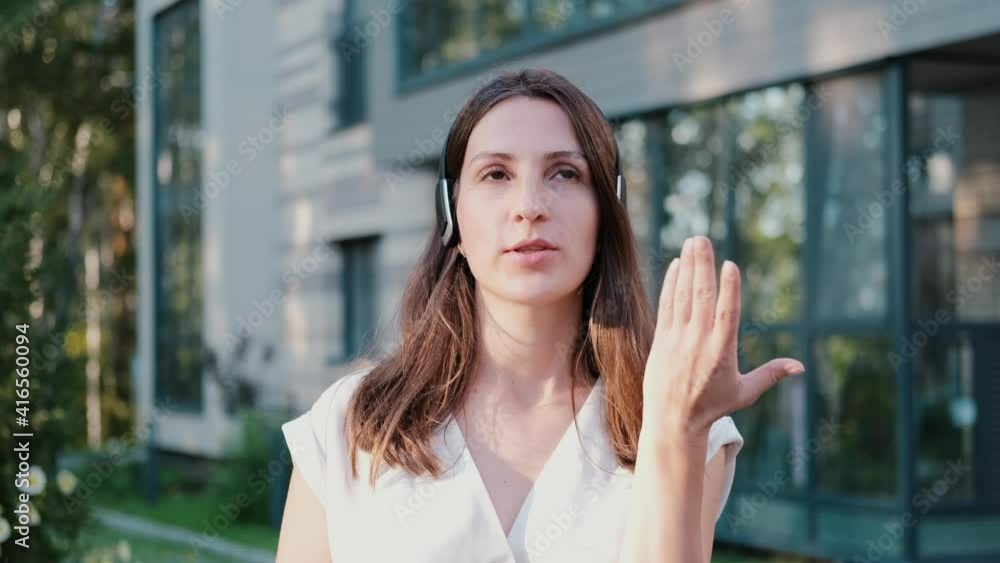 Happy young business woman is wearing headphones, talking to webcam, making remote online videoconference. Female Internet teacher doing remote chat while working outdoors. Webcam view Happy young business woman is wearing headphones, talking to webcam, making remote online videoconference. Female Internet teacher doing remote chat while working outdoors. Webcam view