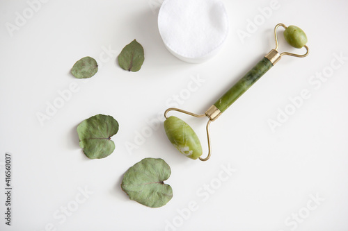 green jade facial massage roller, white cotton pads, eucalyptus petals on white background