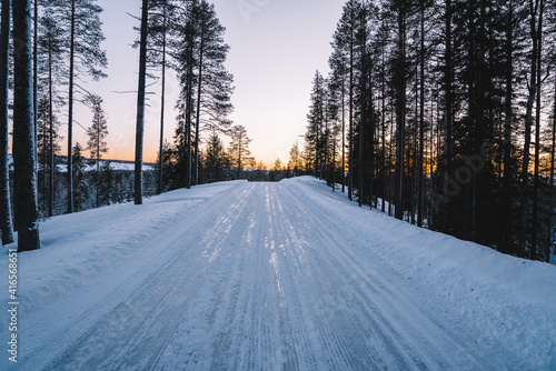 Beautiful natural environment white northern destination with road for traveling, scenic picture of winter season forest with frost and snow. Sunset in the wood between the trees. Selective focus.