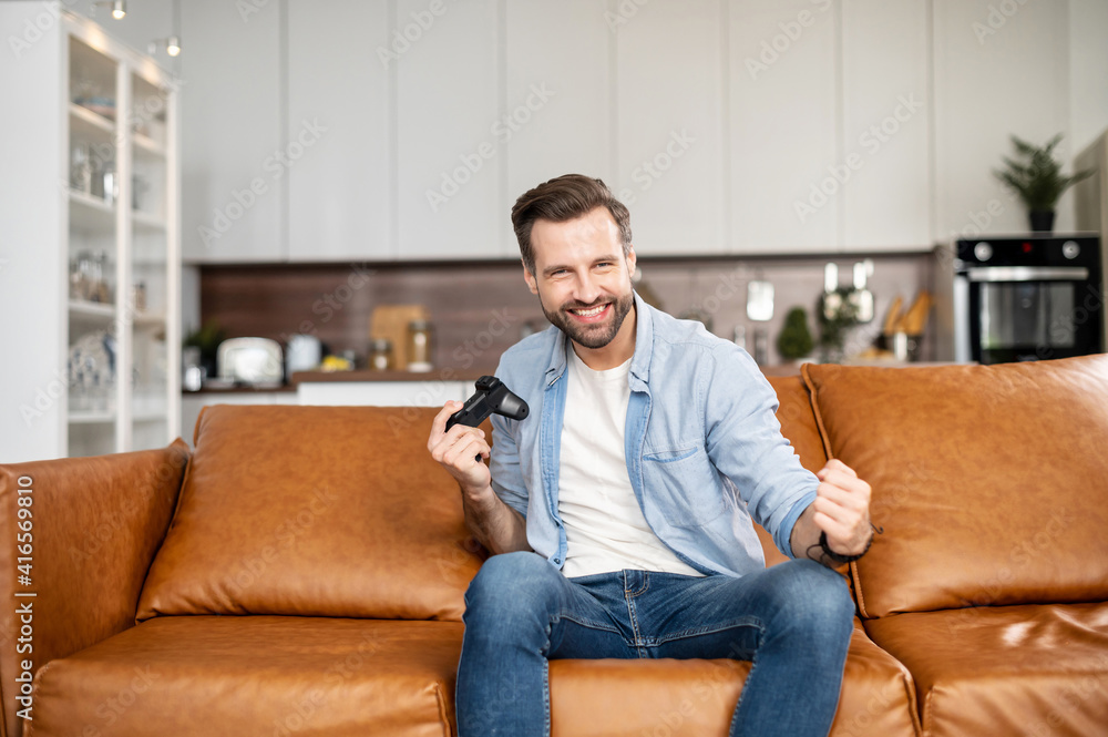 Young bearded domestic man sitting on the couch in the living room staying at home and playing, won a video game match or a battle, holding wireless joystick, making yes gesture, victory concept
