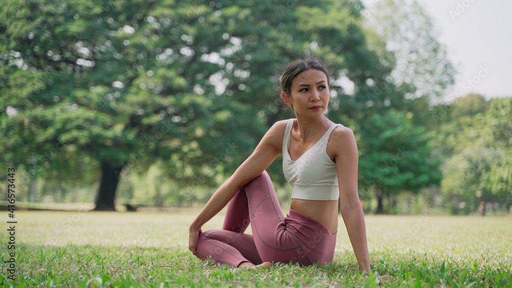Asian young woman sitting on the grass in the lotus position and raising hands up outside in city park with the big trees background Asian young woman sitting on the grass in the lotus position and raising hands up outside in city park with the big trees background