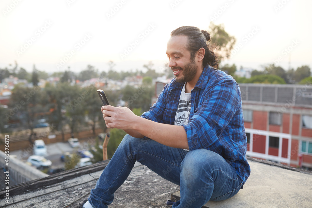 Obraz premium 30 year old man sitting on the roof of a building watching the sunset and 30-year-old man sitting on his back on the rooftop of a building watching the sunset and browsing on his smart cell phone