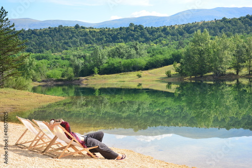 Fototapeta Naklejka Na Ścianę i Meble -  Frau entspannt im Liegestuhl am Idyllischen See Perućko Jezero bei Sinj, Vrlika in Kroatien mit Bäumen und Berge im Hintergrund
