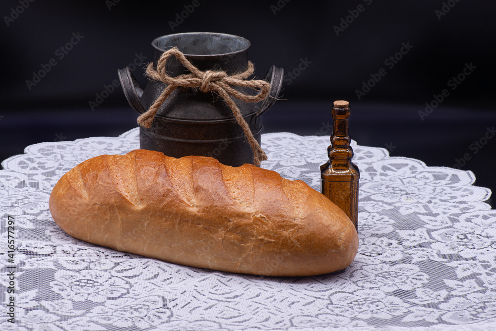 Composition of white bread, small bottle and old jug on white tablecloth on black background
