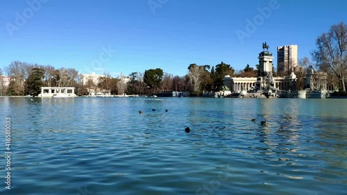 Main pond in the famous Retiro park in Madrid on a sunny winter day. Ducks are simming on the water while seagulls fly around. The monument of Alfonso XII is standing tall to the right of the picture.