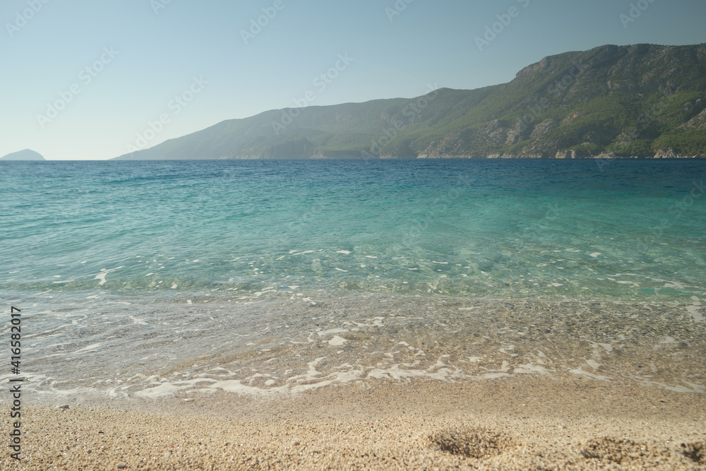 Sandy beach with crystal clear sea water. Green mountains and blue sky in the background. Beautiful marine landscape.