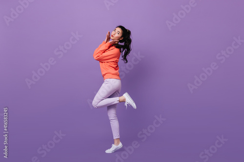 Charming asian girl with ponytail standing on one leg. Full length view of amazing japanese model dancing on purple background.