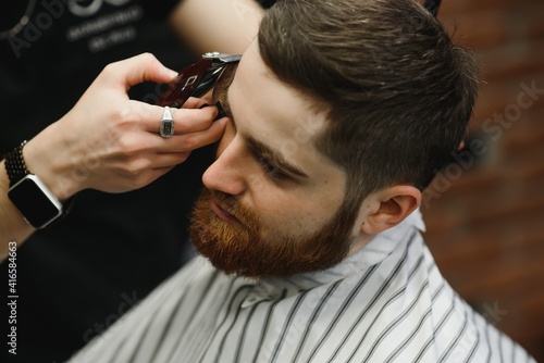 Making haircut look perfect. Young bearded man getting haircut by hairdresser while sitting in chair at barbershop