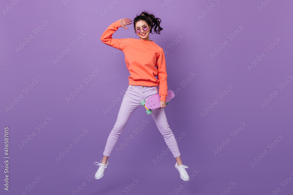 Funny asian girl in pants jumping on purple background. Full length view of korean young woman with longboard.