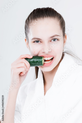 Beautiful girl in a white robe bites a cucumber looking at the camera on a light background