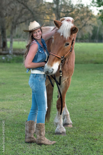Young woman in jeans, boots and cowgirl hat smiling with her horse