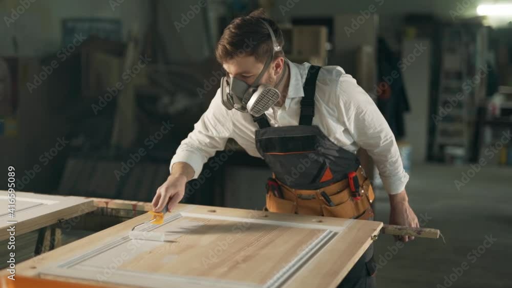 Man in work uniform and respirators paints a wooden product with a roller in white Man in work uniform and respirators paints a wooden product with a roller in white