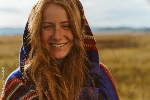 Against the background of the steppe in a bright beautiful cape on her head in ethno style smiles a beautiful smile girl with white teeth and red long hair. High quality photo