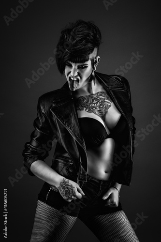 Pretty young tattooed woman, full face, with punk hairstyle, wearing bra, leather jacket, fishnet stockings, sticking out her tongue