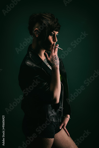 Attractive tattooed young woman, in profile, with punk hairstyle, wearing leather jacket, fishnet stockings, smoking a cigarette © Warpedgalerie
