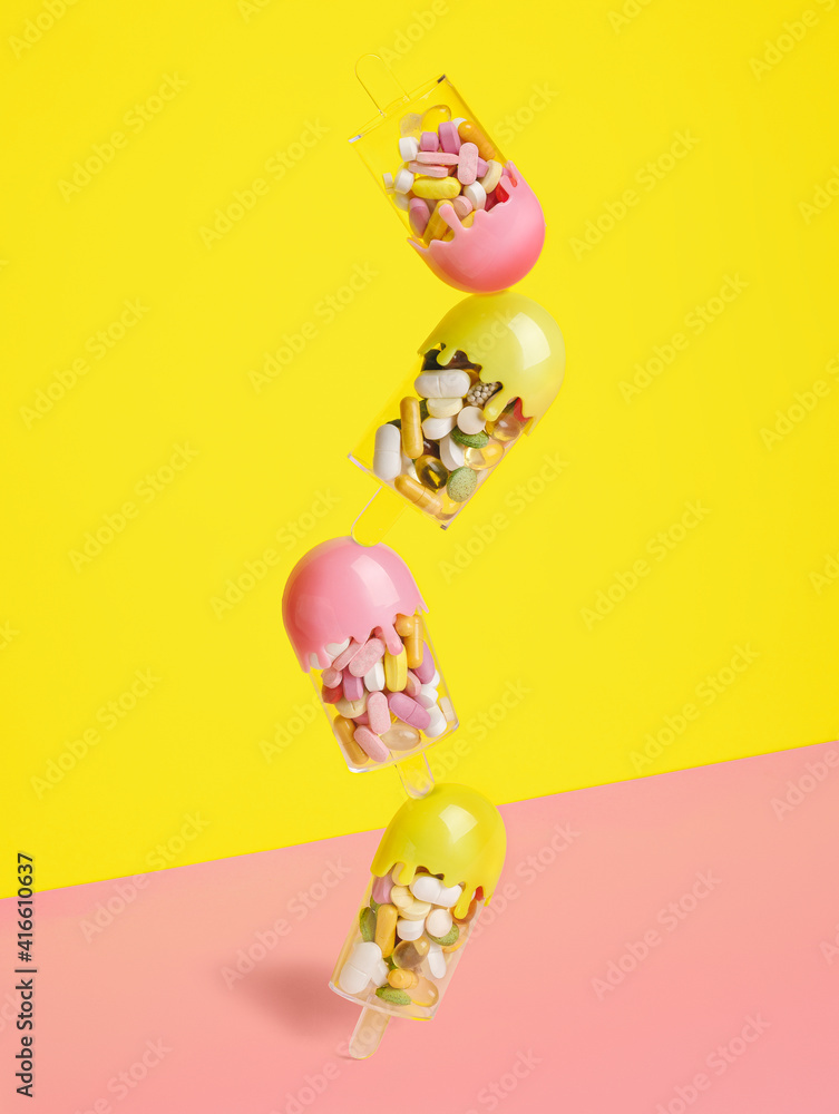 Vertically assorted popsicle ice cream filled with various pills and tablets on two tone pink and yellow background. Creative health care or summer sweet food concept. Minimal flat lay.