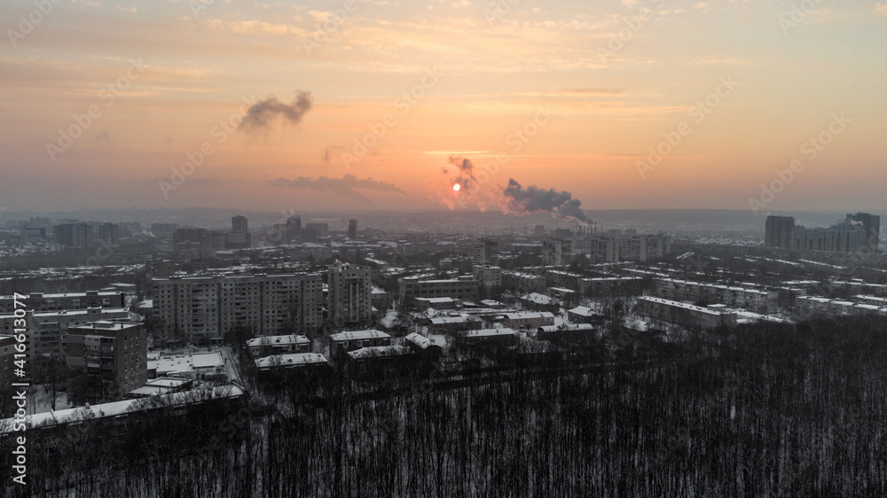 Aerial view Kharkiv city Pavlove Pole district. Smoke from pipes above winter dark forest, multistorey houses and snowy orange sunset sky