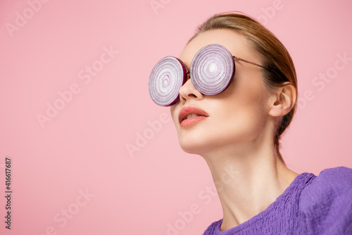 Fototapeta Naklejka Na Ścianę i Meble -  young woman posing in eyeglasses with purple onion rings isolated on pink, surrealism concept