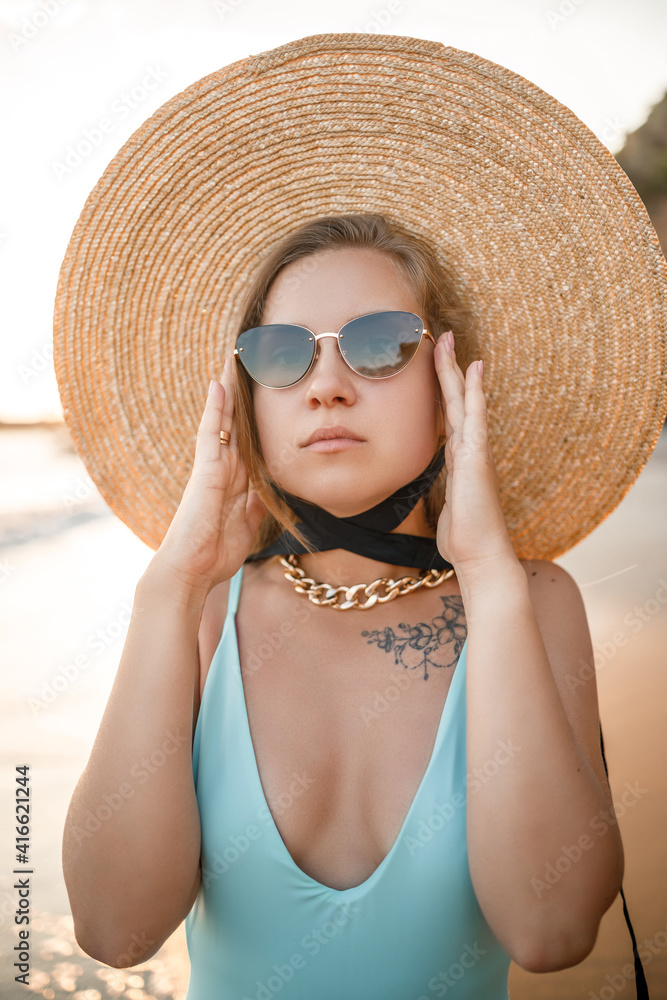 A young tanned woman in a beautiful swimsuit with a straw hat stands and rests on a tropical beach with sand and looks at the sunset and the sea. Selective focus. Vacation concept by the sea