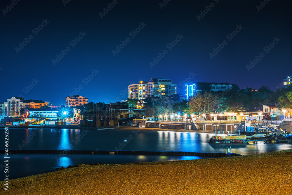 Gelendzhik sea resort beach coastline at night with illuminated modern buildings at background reflected in sea surface .