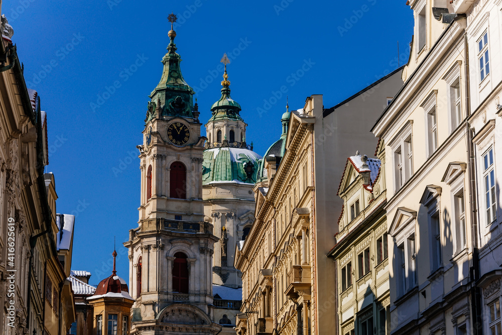 View of Baroque Church of Saint Nicholas, green dome and bell tower with clock, sunny winter day, snow on red roofs, Mala Strana or Lesser Town district, Prague, Czech Republic