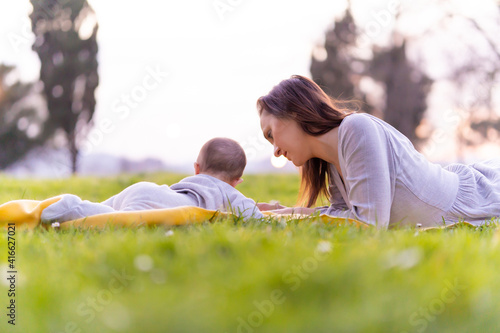 beautiful middle-aged young woman lying in a park in gray dress with her six-month-old baby at sunset in spring or summer. maternity lifestyle concept