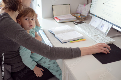 Mother multi-tasking, holding girl kid and using computer at home. Candid authentic and real life mom working and parenting.