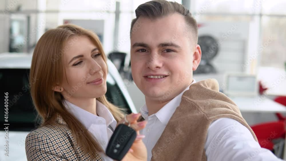 Happy couple taking a selfie at a car dealership with a new car in the background. Man and woman showing keys of his new car in dealership Happy couple taking a selfie at a car dealership with a new car in the background. Man and woman showing keys of his new car in dealership