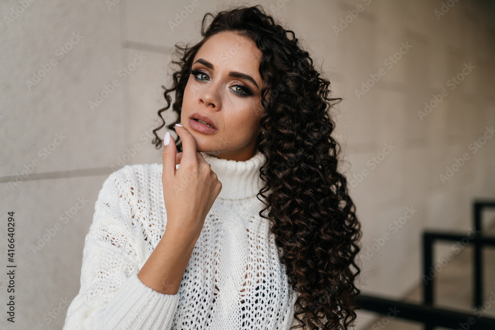 Against the grey wall close-up of a girl with beautiful facial features with a thick mane of curly dark hair touching her chin with her hand very gently. High quality photo Against the grey wall close-up of a girl with beautiful facial features with a thick mane of curly dark hair touching her chin with her hand very gently. High quality photo